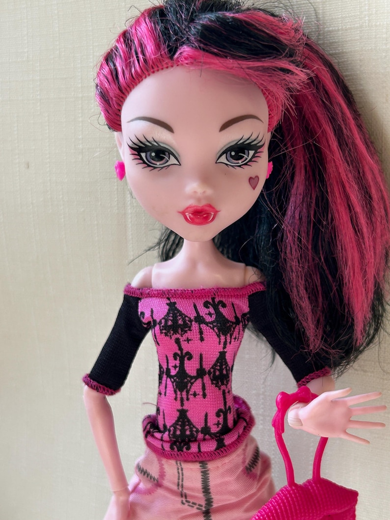 May include: A doll with pink and black hair, wearing a pink and black top and a pink skirt. The doll has pink earrings, a heart-shaped mark on its cheek, and is holding a pink purse. The doll's lips are red.