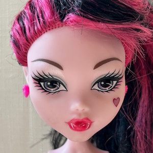 May include: A close-up of a doll's face with pink and black hair. The doll has pale skin, large eyes with long eyelashes, and red lips. A small heart is painted on the cheek. The doll is wearing pink earrings.