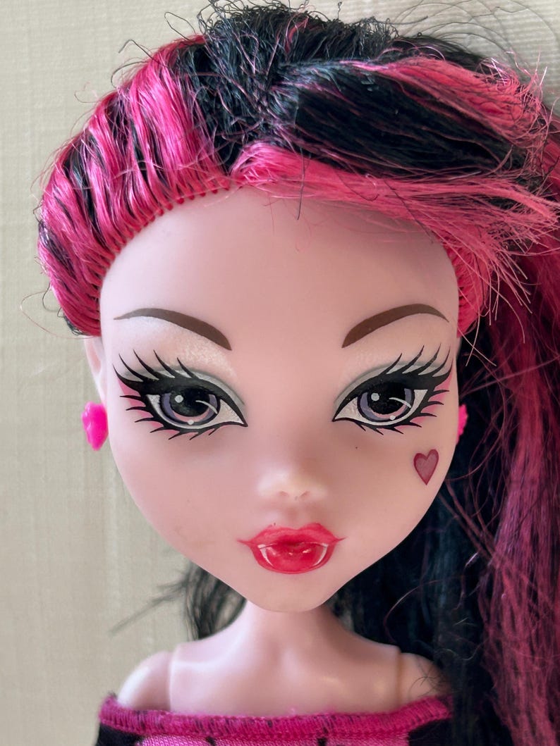 May include: A close-up of a doll's face with pink and black hair. The doll has pale skin, large eyes with long eyelashes, and red lips. A small heart is painted on the cheek. The doll is wearing pink earrings.
