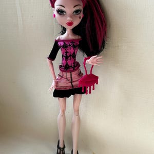May include: A doll with black and pink hair, wearing a pink and black outfit, black shoes, and holding a red purse. The doll has pale skin, red lips, and a heart-shaped mark on her cheek. The doll's outfit includes a pink skirt and a black top.