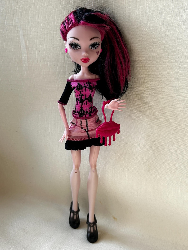 May include: A doll with black and pink hair, wearing a pink and black outfit, black shoes, and holding a red purse. The doll has pale skin, red lips, and a heart-shaped mark on her cheek. The doll's outfit includes a pink skirt and a black top.