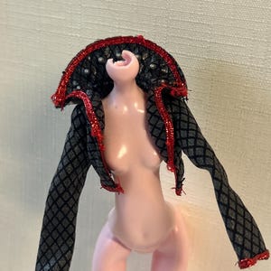 Puede incluir: Una mu&ntilde;eca con una chaqueta estampada en negro y gris con ribete rojo. La chaqueta tiene un cuello alto con tachuelas plateadas y ribete rojo. La mu&ntilde;eca est&aacute; desnuda con piel rosada.