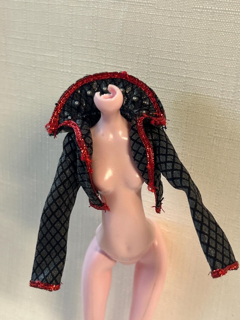 Puede incluir: Una mu&ntilde;eca con una chaqueta estampada en negro y gris con ribete rojo. La chaqueta tiene un cuello alto con tachuelas plateadas y ribete rojo. La mu&ntilde;eca est&aacute; desnuda con piel rosada.