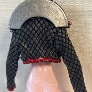 Puede incluir: Una chaqueta de mu&ntilde;eca negra y gris con un patr&oacute;n de diamantes, ribete rojo y cuello plateado. El cuello plateado tiene un dise&ntilde;o de murci&eacute;lago y telara&ntilde;a. La chaqueta est&aacute; sobre una mu&ntilde;eca rosa.