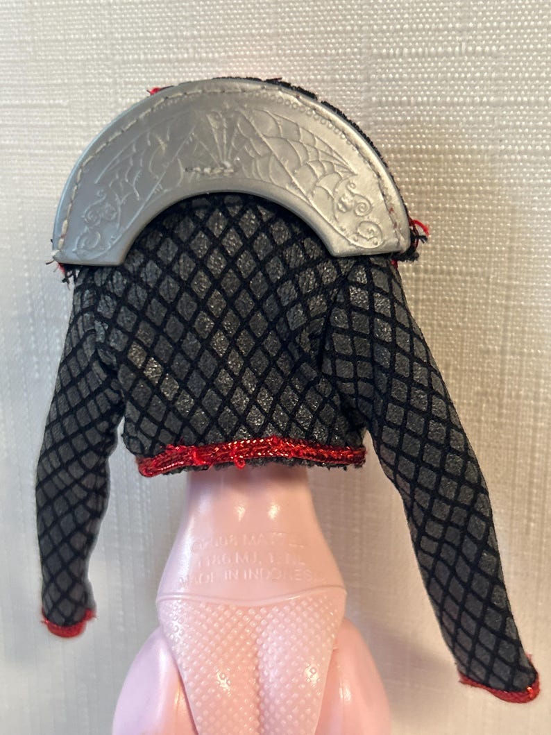 Puede incluir: Una chaqueta de mu&ntilde;eca negra y gris con un patr&oacute;n de diamantes, ribete rojo y cuello plateado. El cuello plateado tiene un dise&ntilde;o de murci&eacute;lago y telara&ntilde;a. La chaqueta est&aacute; sobre una mu&ntilde;eca rosa.