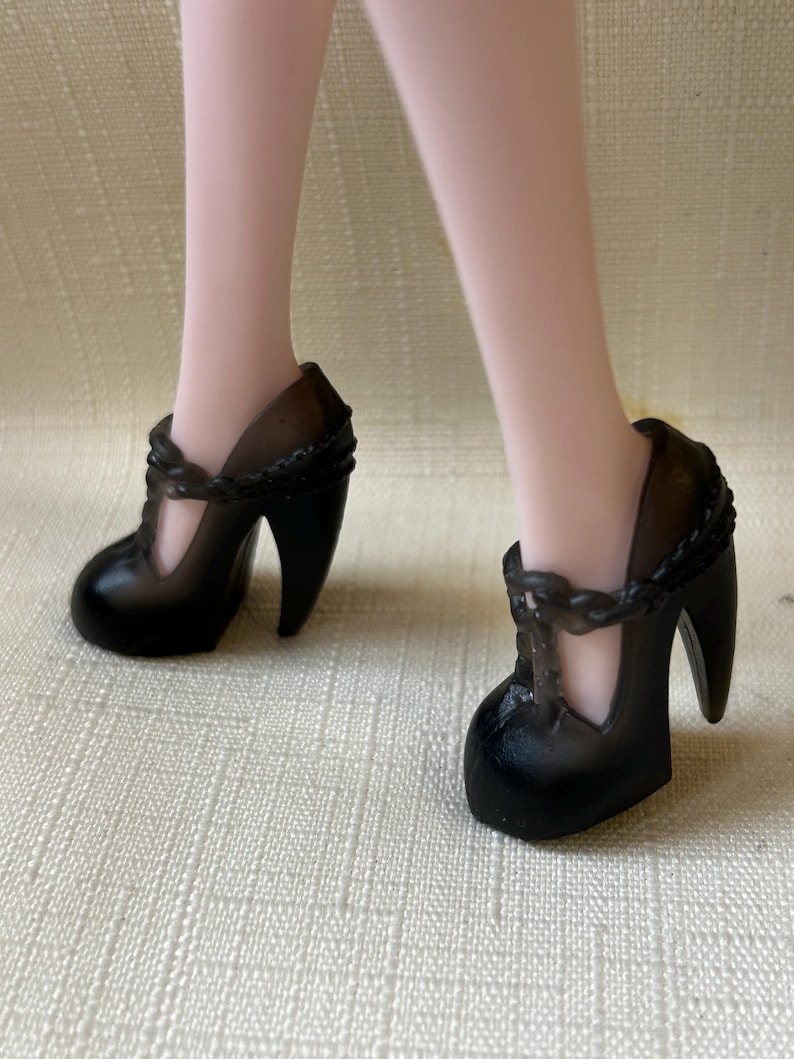 May include: Black high-heeled shoes with a T-strap design. The shoes have a rounded toe and a thick heel. The T-strap is adorned with a braided detail. The shoes are on a light-colored surface.