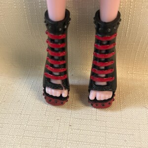 Puede incluir: Sandalias negras y rojas de tac&oacute;n alto con dise&ntilde;o de tiras. Las sandalias tienen un ribete tachonado y las lleva una mu&ntilde;eca. Las sandalias tienen un dise&ntilde;o de punta abierta.