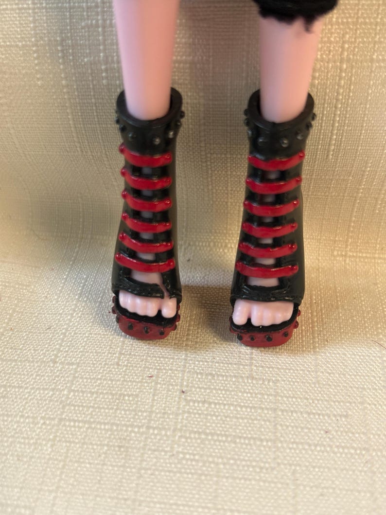 Puede incluir: Sandalias negras y rojas de tac&oacute;n alto con dise&ntilde;o de tiras. Las sandalias tienen un ribete tachonado y las lleva una mu&ntilde;eca. Las sandalias tienen un dise&ntilde;o de punta abierta.