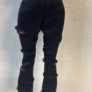 Puede incluir: Pantalones negros de mu&ntilde;eca con detalles desgastados. Los pantalones tienen m&uacute;ltiples rasgaduras y desgarros a lo largo de las piernas, revelando una tela roja debajo. Los pantalones est&aacute;n hechos de un material el&aacute;stico.