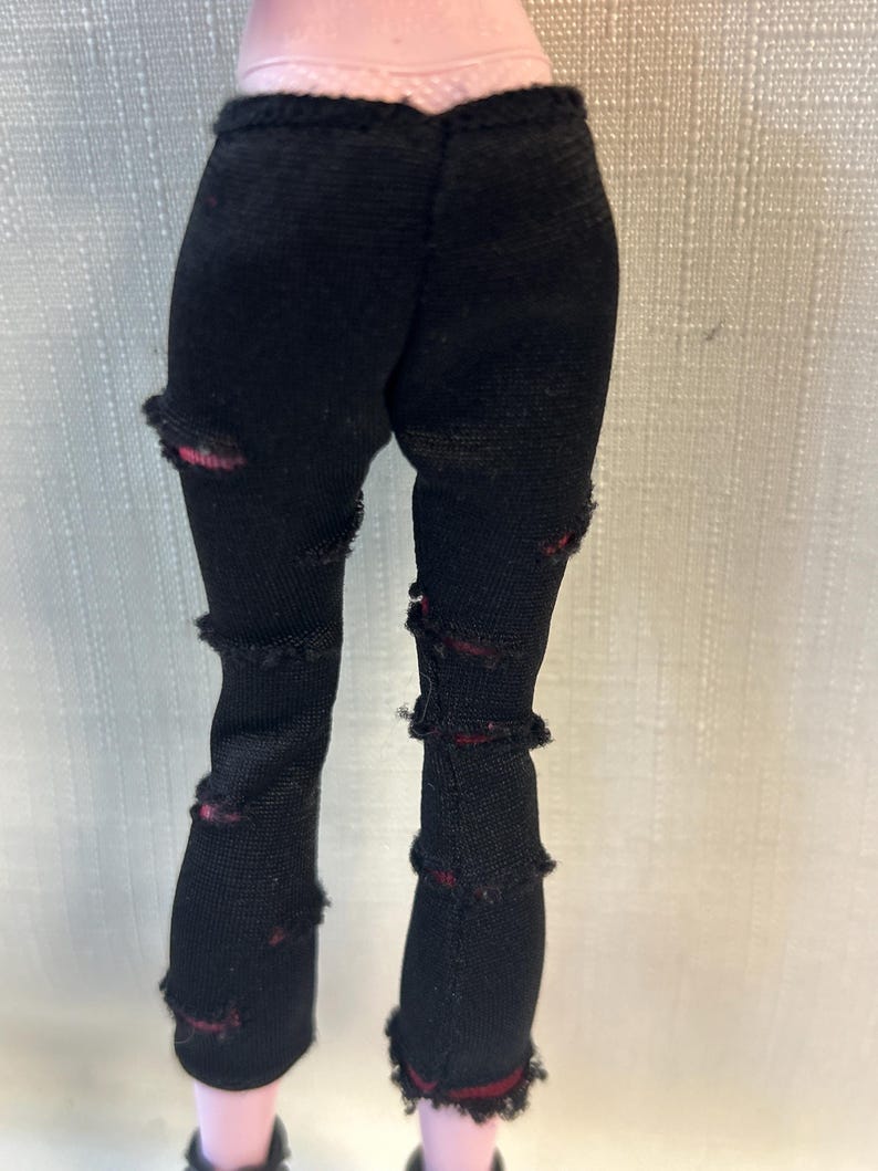 Puede incluir: Pantalones negros de mu&ntilde;eca con detalles desgastados. Los pantalones tienen m&uacute;ltiples rasgaduras y desgarros a lo largo de las piernas, revelando una tela roja debajo. Los pantalones est&aacute;n hechos de un material el&aacute;stico.