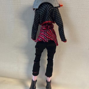 Puede incluir: Una mu&ntilde;eca vestida con un atuendo negro y rojo. La mu&ntilde;eca tiene pantalones negros con rasgaduras, una blusa a rayas rojas y negras y una chaqueta negra con un patr&oacute;n de diamantes grises. La mu&ntilde;eca lleva botas negras con tacones rojos.