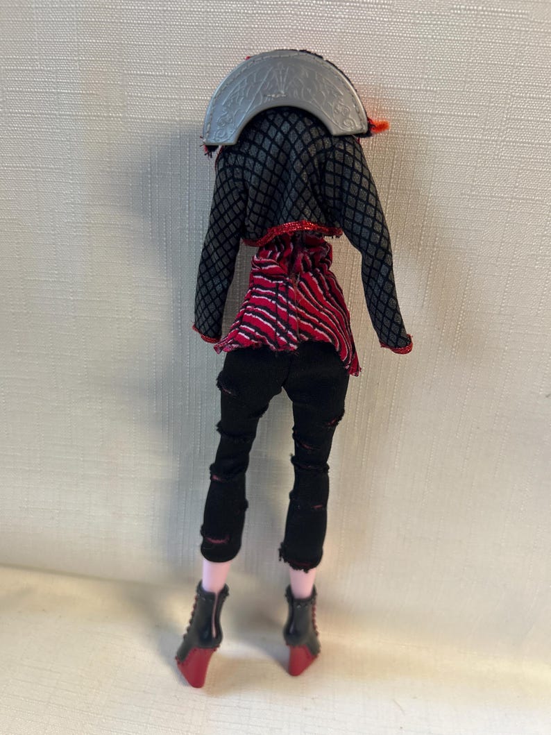 Puede incluir: Una mu&ntilde;eca vestida con un atuendo negro y rojo. La mu&ntilde;eca tiene pantalones negros con rasgaduras, una blusa a rayas rojas y negras y una chaqueta negra con un patr&oacute;n de diamantes grises. La mu&ntilde;eca lleva botas negras con tacones rojos.