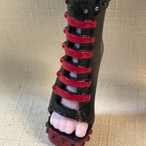 Puede incluir: Zapato de mu&ntilde;eca negro y rojo con tac&oacute;n alto. El zapato presenta una base negra con detalles rojos y una serie de correas rojas. El tac&oacute;n est&aacute; tachonado con peque&ntilde;os adornos plateados. Se ve el pie de la mu&ntilde;eca.
