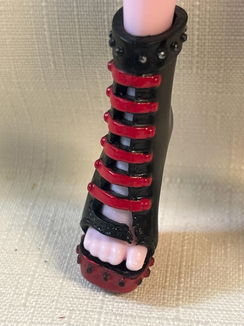 Puede incluir: Zapato de mu&ntilde;eca negro y rojo con tac&oacute;n alto. El zapato presenta una base negra con detalles rojos y una serie de correas rojas. El tac&oacute;n est&aacute; tachonado con peque&ntilde;os adornos plateados. Se ve el pie de la mu&ntilde;eca.