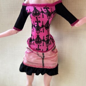 May include: A doll wearing a pink and black dress. The off-the-shoulder top is pink with a black pattern and black sleeves. The skirt is pink with a black ruffled hem. The doll has pink skin and black and pink hair.