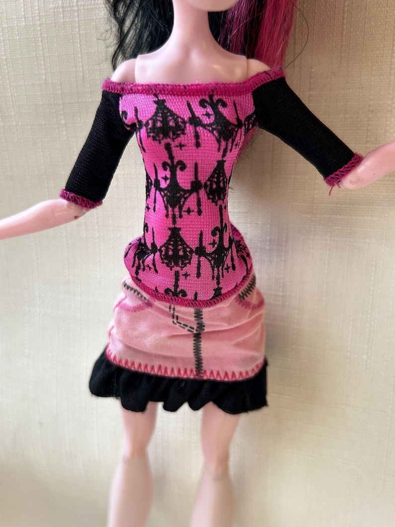 May include: A doll wearing a pink and black dress. The off-the-shoulder top is pink with a black pattern and black sleeves. The skirt is pink with a black ruffled hem. The doll has pink skin and black and pink hair.