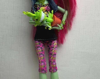 May include: A Monster High doll with green skin, pink hair, and a black top. She wears patterned pink and yellow leggings and purple boots. The doll holds a green frog-like creature in an orange pot. The doll has a denim jacket.