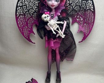 May include: A Monster High doll with pink and black wings, holding a small skeleton. The doll has pink hair and is wearing a pink and black dress. Accessories include a pink mask and a pink container.