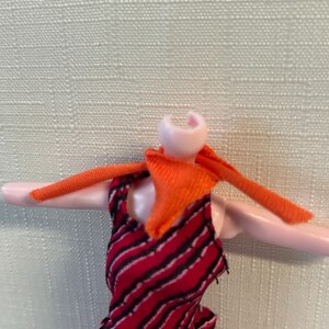 Puede incluir: Una mu&ntilde;eca con un vestido a rayas rojas y negras con una bufanda naranja. Los brazos de la mu&ntilde;eca est&aacute;n extendidos y el vestido tiene un dobladillo con volantes. Falta la cabeza de la mu&ntilde;eca.