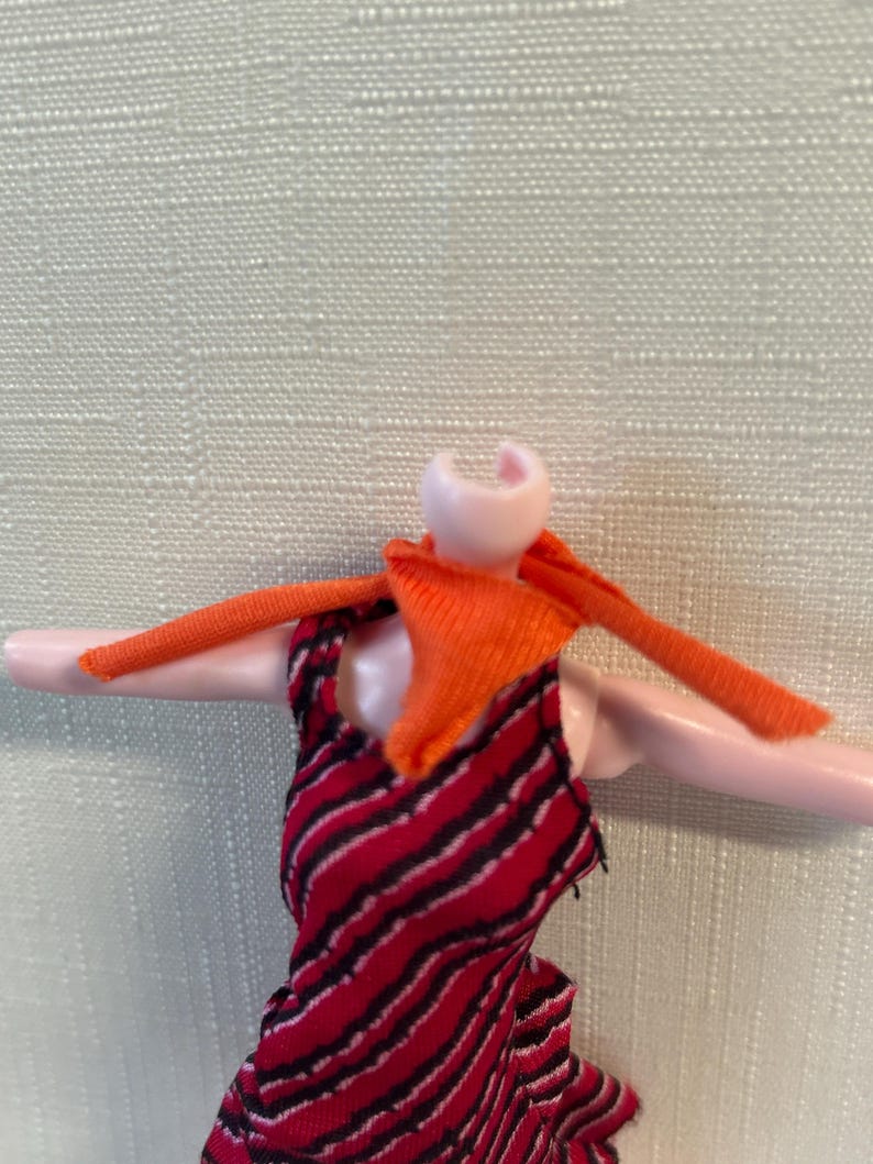 Puede incluir: Una mu&ntilde;eca con un vestido a rayas rojas y negras con una bufanda naranja. Los brazos de la mu&ntilde;eca est&aacute;n extendidos y el vestido tiene un dobladillo con volantes. Falta la cabeza de la mu&ntilde;eca.