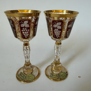 May include: Two matching crystal goblets featuring deep red panels with etched white grape vine motifs. The rims and bases are trimmed in gold, and the stems have a clear, textured design. Each base displays a gold Egermann sticker.
