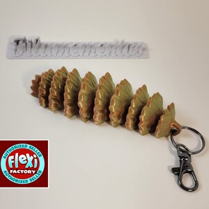 May include: A gold and green 3D printed pine cone keychain with a black metal key ring. The keychain is attached to a white label with the text "Filamentous".