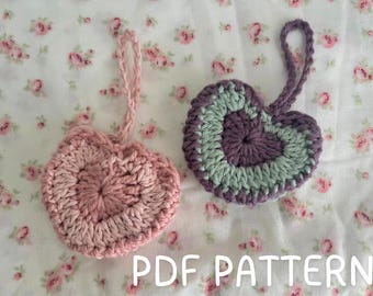 Peut inclure: Deux ornements en forme de cœur en crochet, l'un en rose et l'autre en violet et vert. Les ornements sont suspendus sur un fond blanc à motif floral. Le texte "PDF PATTERN" est visible en bas de l'image.