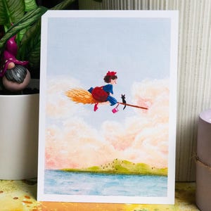 May include: A whimsical illustration depicts a young witch in a blue dress, red bow, and pink shoes, riding a broom with a black cat. The artwork is set against a backdrop of fluffy clouds, a blue sky, and a serene landscape.