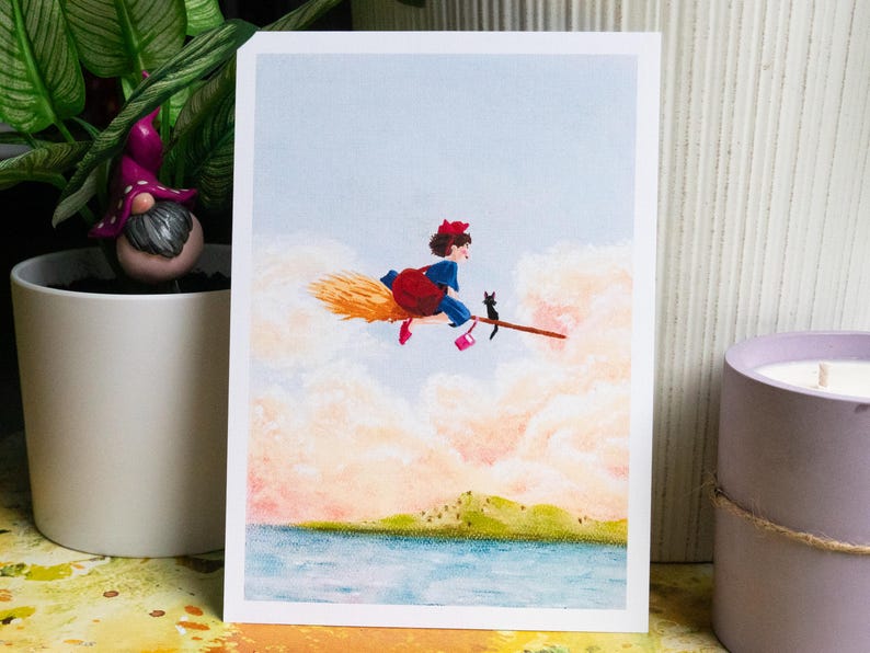 May include: A whimsical illustration depicts a young witch in a blue dress, red bow, and pink shoes, riding a broom with a black cat. The artwork is set against a backdrop of fluffy clouds, a blue sky, and a serene landscape.