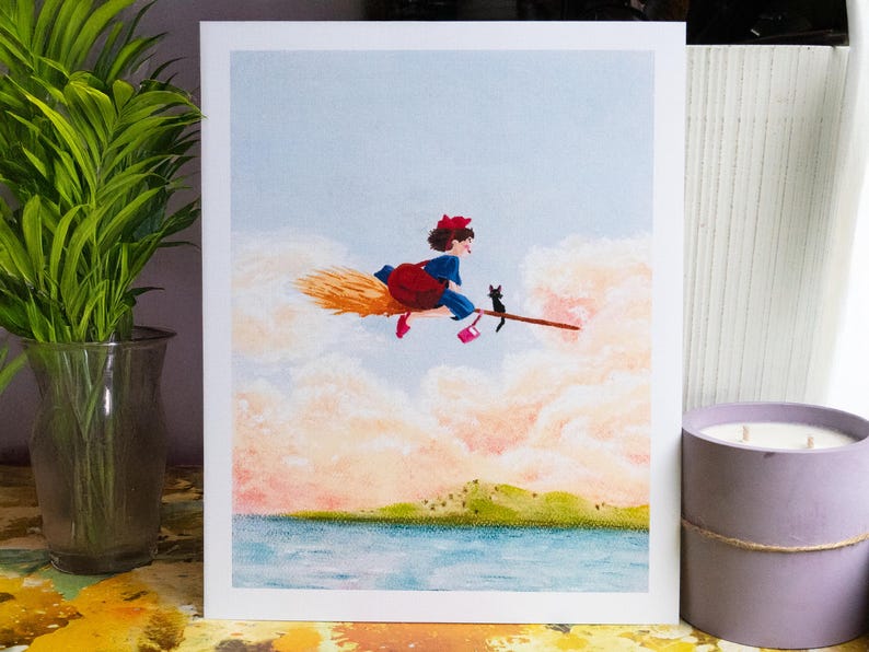 May include: A whimsical illustration depicts a young witch in a blue dress and red bow riding a broom with a black cat. The artwork is set against a backdrop of fluffy clouds, a blue sky, and a body of water.