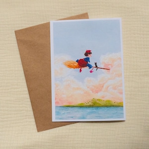 Puede incluir: Tarjeta de felicitaci&oacute;n con una ilustraci&oacute;n caprichosa de una bruja volando en una escoba con un gato negro. La bruja lleva un lazo rojo y un vestido azul, volando sobre un paisaje con un mar azul y nubes rosas. Incluye un sobre marr&oacute;n.