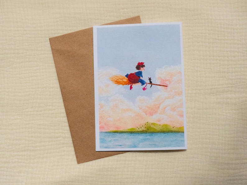 Puede incluir: Tarjeta de felicitaci&oacute;n con una ilustraci&oacute;n caprichosa de una bruja volando en una escoba con un gato negro. La bruja lleva un lazo rojo y un vestido azul, volando sobre un paisaje con un mar azul y nubes rosas. Incluye un sobre marr&oacute;n.
