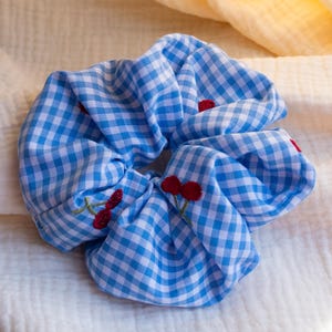 Könnte beinhalten: Ein blau-weiß kariertes Haargummi mit gestickten roten Kirschen. Das Haaraccessoire ist aus Stoff gefertigt und soll das Haar an Ort und Stelle halten. Das Haargummi ist ein stilvolles und funktionelles Haaraccessoire.
