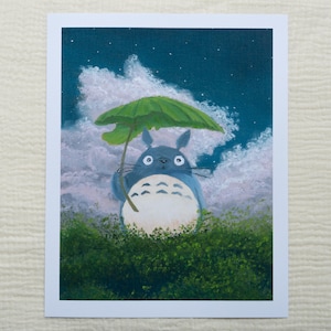 May include: A print of Totoro holding a leaf umbrella, set against a night sky with clouds and stars. The character is blue with a white belly, standing in green foliage. The artwork is framed in white.