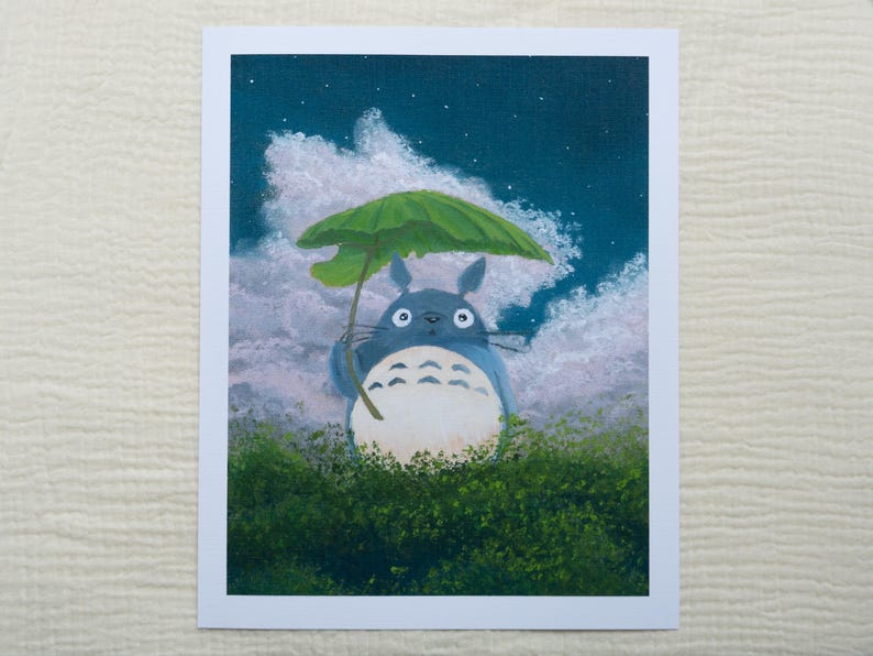 May include: A print of Totoro holding a leaf umbrella, set against a night sky with clouds and stars. The character is blue with a white belly, standing in green foliage. The artwork is framed in white.