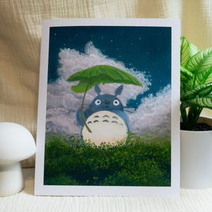 May include: A print of Totoro holding a green umbrella in a night scene with clouds and stars. The artwork features a blue Totoro character standing in green foliage. A white mushroom-shaped decor is in the foreground.