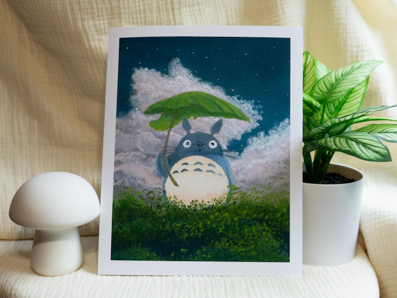 May include: A print of Totoro holding a green umbrella in a night scene with clouds and stars. The artwork features a blue Totoro character standing in green foliage. A white mushroom-shaped decor is in the foreground.