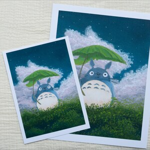 May include: Two art prints featuring a blue Totoro character holding a green leaf umbrella. The background shows a night sky with clouds and stars. The prints are on white paper and set against a textured white surface.