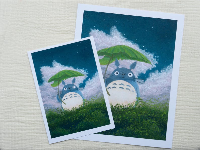May include: Two art prints featuring a blue Totoro character holding a green leaf umbrella. The background shows a night sky with clouds and stars. The prints are on white paper and set against a textured white surface.