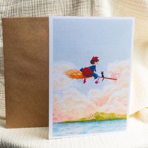 Puede incluir: Tarjeta de felicitaci&oacute;n con una ilustraci&oacute;n de una joven bruja en una escoba con un gato negro, sobre un fondo de cielo pastel y nubes. Una envoltura marr&oacute;n est&aacute; al lado de la tarjeta. La obra de arte representa una masa de agua y un paisaje verde.