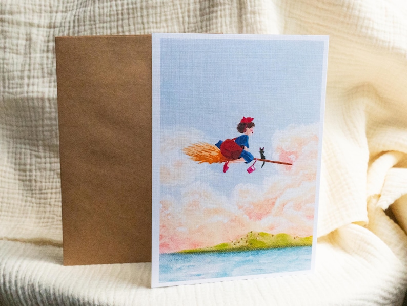 Puede incluir: Tarjeta de felicitaci&oacute;n con una ilustraci&oacute;n de una joven bruja en una escoba con un gato negro, sobre un fondo de cielo pastel y nubes. Una envoltura marr&oacute;n est&aacute; al lado de la tarjeta. La obra de arte representa una masa de agua y un paisaje verde.