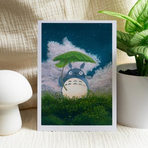 May include: A print featuring a blue Totoro character holding a green umbrella, set against a cloudy night sky. The artwork is framed with a white border and set on a white surface. A white mushroom-shaped decor and a potted plant are also in the image.