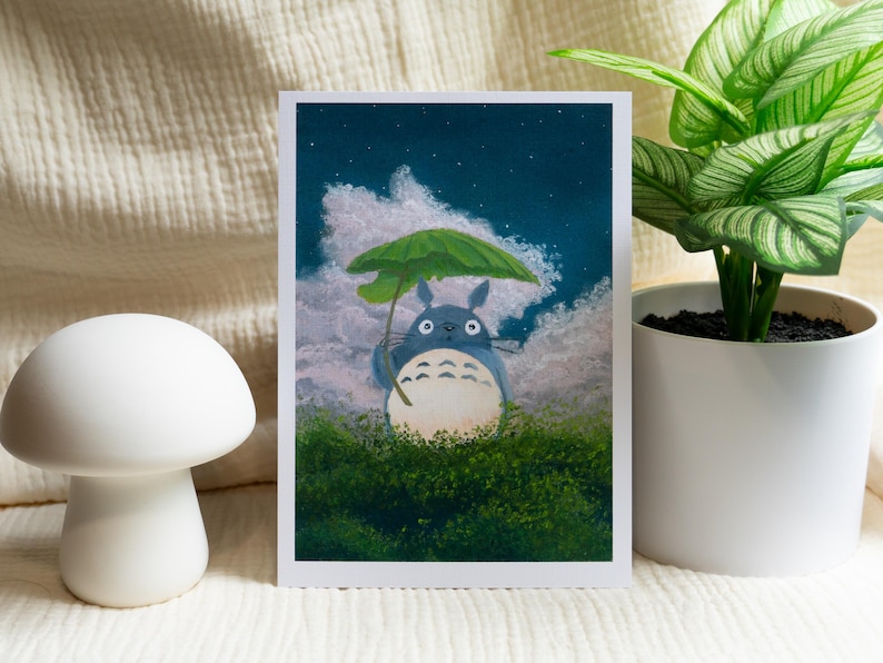 May include: A print featuring a blue Totoro character holding a green umbrella, set against a cloudy night sky. The artwork is framed with a white border and set on a white surface. A white mushroom-shaped decor and a potted plant are also in the image.