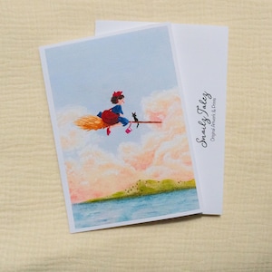 Puede incluir: Tarjeta de felicitaci&oacute;n con una ilustraci&oacute;n de una joven bruja volando en una escoba con un gato negro. La obra de arte est&aacute; ambientada sobre un fondo de cielo azul y nubes esponjosas. Incluye el texto "Snaily Talez Original Artwork & Prints".