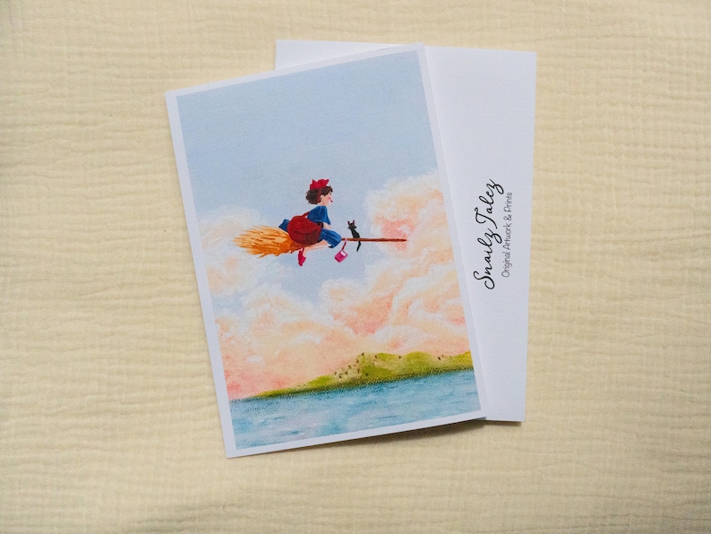 Puede incluir: Tarjeta de felicitaci&oacute;n con una ilustraci&oacute;n de una joven bruja volando en una escoba con un gato negro. La obra de arte est&aacute; ambientada sobre un fondo de cielo azul y nubes esponjosas. Incluye el texto "Snaily Talez Original Artwork & Prints".