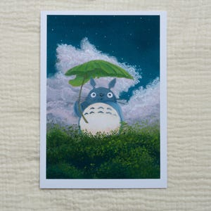 May include: A print of Totoro holding a green umbrella, standing in a field of green bushes. The background features a night sky with clouds and stars. The artwork is on a white rectangular card.