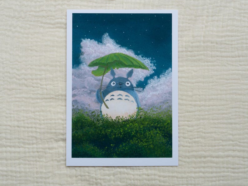 May include: A print of Totoro holding a green umbrella, standing in a field of green bushes. The background features a night sky with clouds and stars. The artwork is on a white rectangular card.
