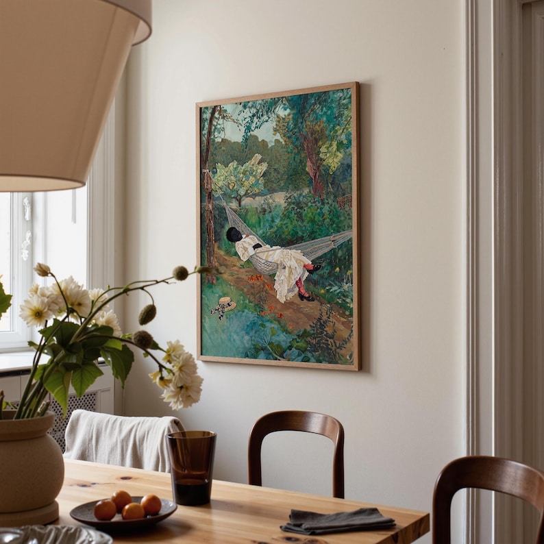 May include: A framed art print depicts a person relaxing in a hammock in a lush forest. The artwork features a person in white clothing, black shoes, and a black hat. The print is in a wooden frame and hangs on a white wall.