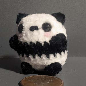 May include: A small, white and black plush panda bear with large black eyes. The panda is standing on a penny.