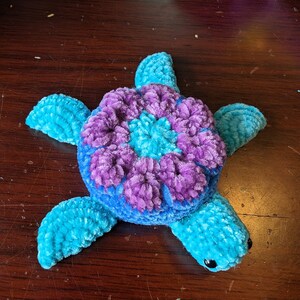 May include: A crocheted stuffed sea turtle toy with a blue and purple shell. The shell has a flower design in the center.